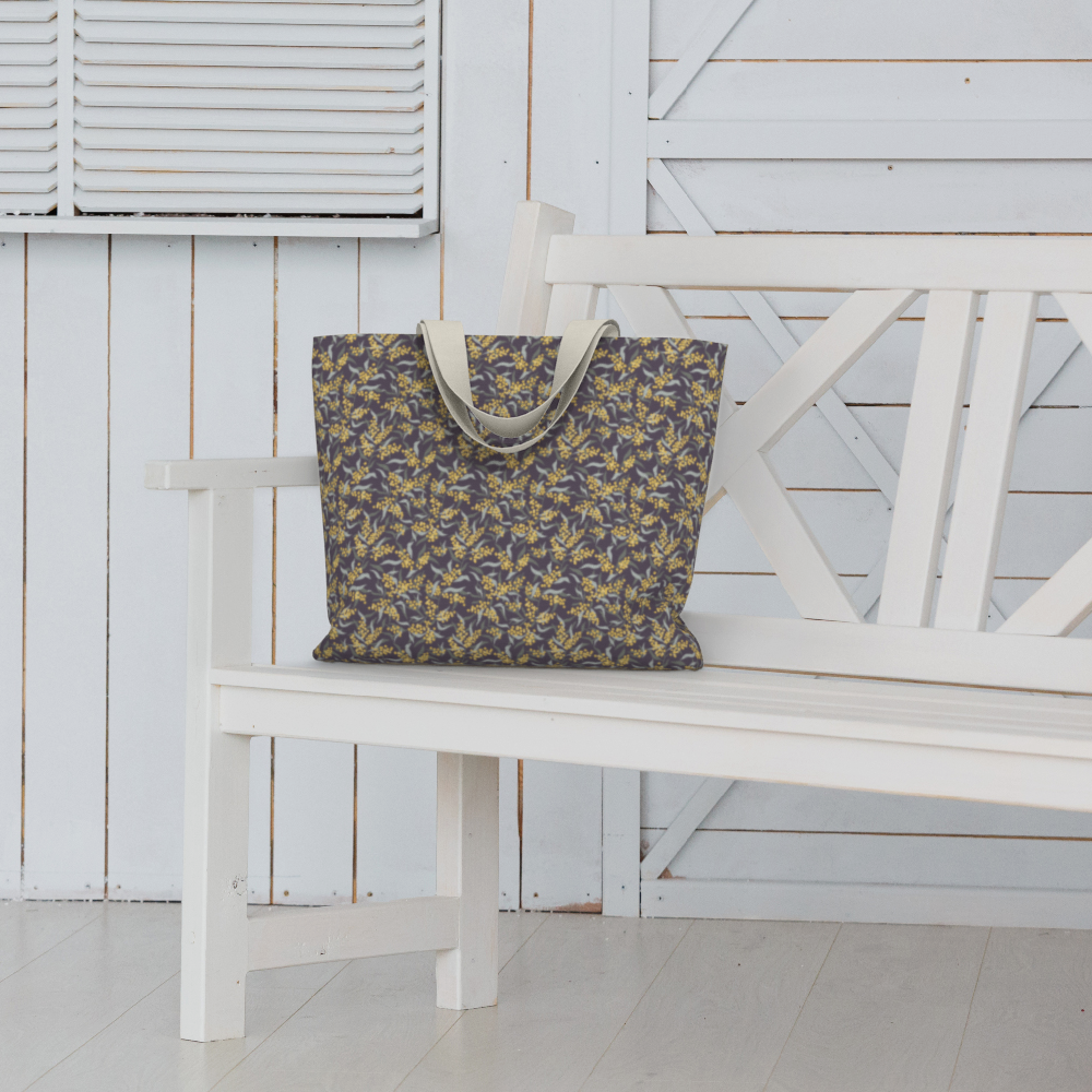 Oversized Fully Lined Cotton Tote Bag with sturdy 1 ½ inch strap. Floral botanical pattern featuring sage green leaves and yellow blossoms on a muted purple background. Shown on white bench.