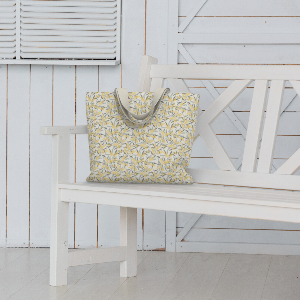 Oversized Fully Lined Cotton Tote Bag with sturdy 1 ½ inch strap. Floral botanical pattern featuring sage green leaves and yellow blossoms on a creamy white background. Shown on white bench.
